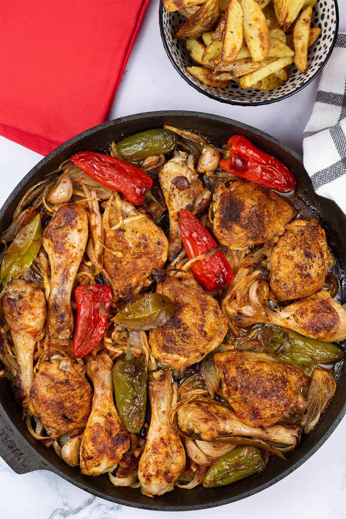 Paprika roast chicken in a large frying pan with a bowl of chunky oven chips and red napkin in the background.