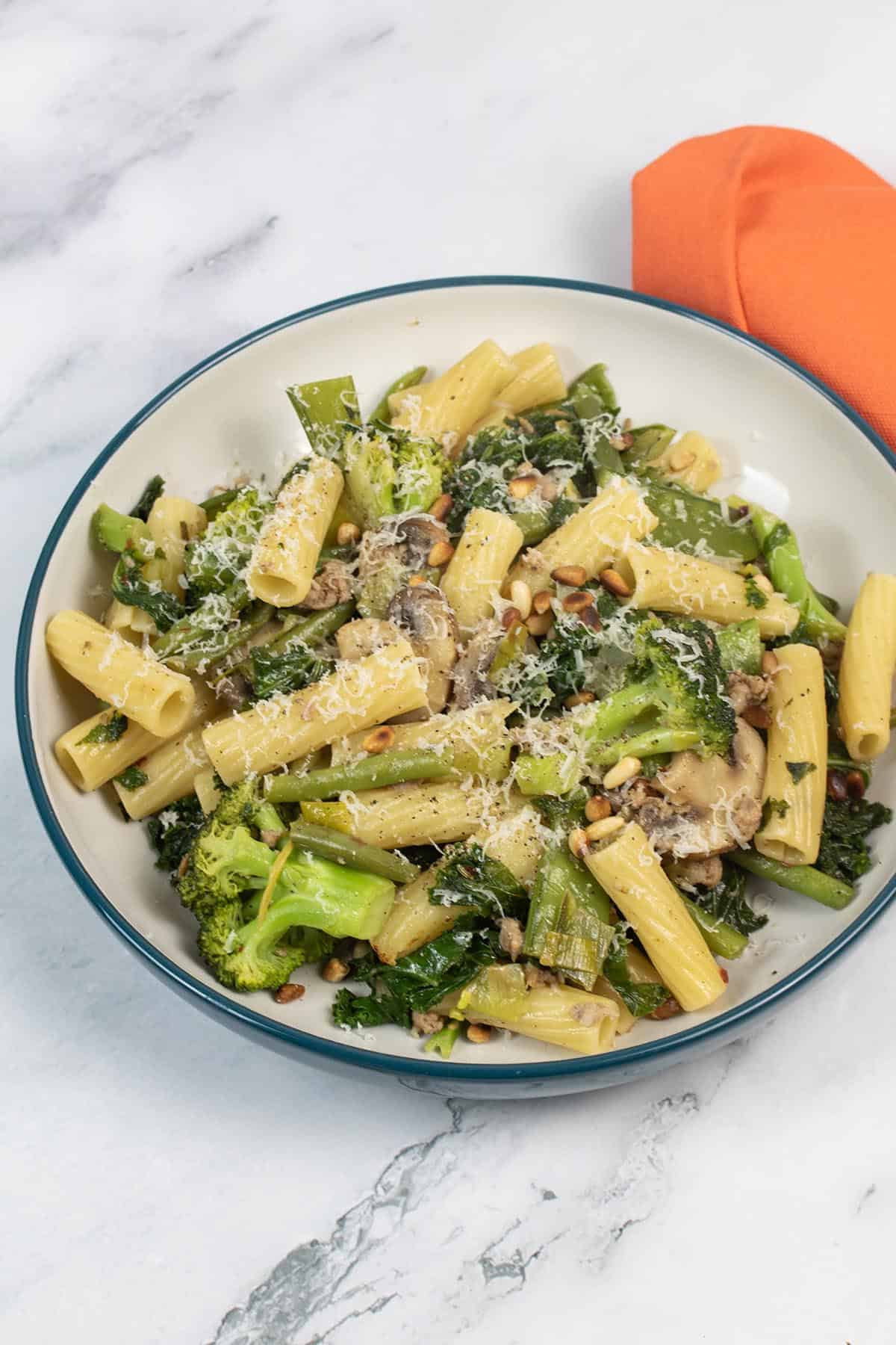 Sausage, vegetable and pine nut pasta in a bowl with an orange napkin on the side.
