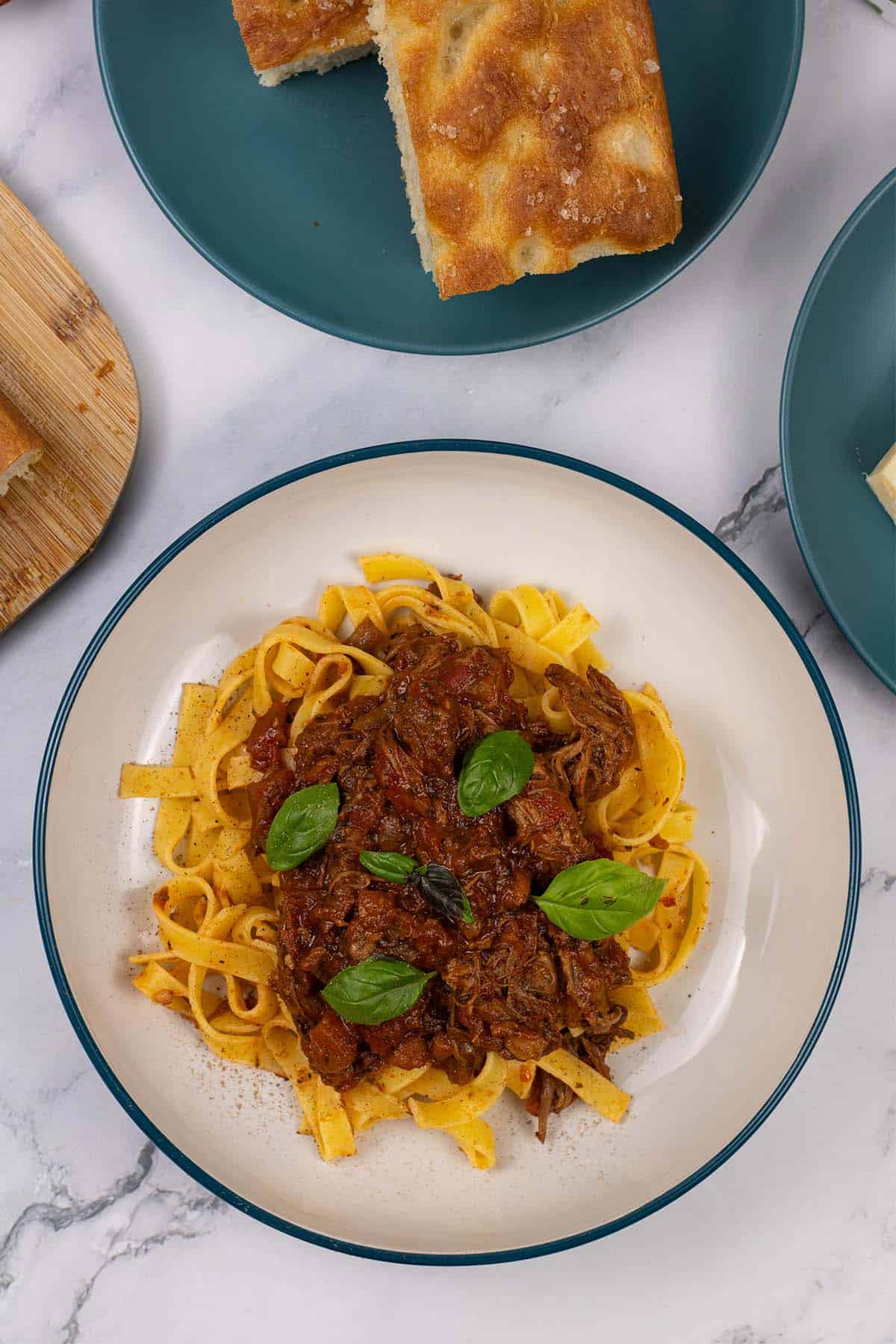 A portion of slow cooker beef ragu in a pasta bowl with tagliatelle and a teal side plate with slices of focaccia in the background.