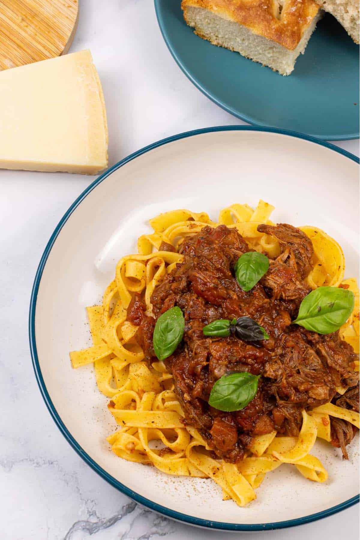 A portion of slow cooker beef ragu in a pasta bowl with tagliatelle and a teal side plate with slices of focaccia in the background.