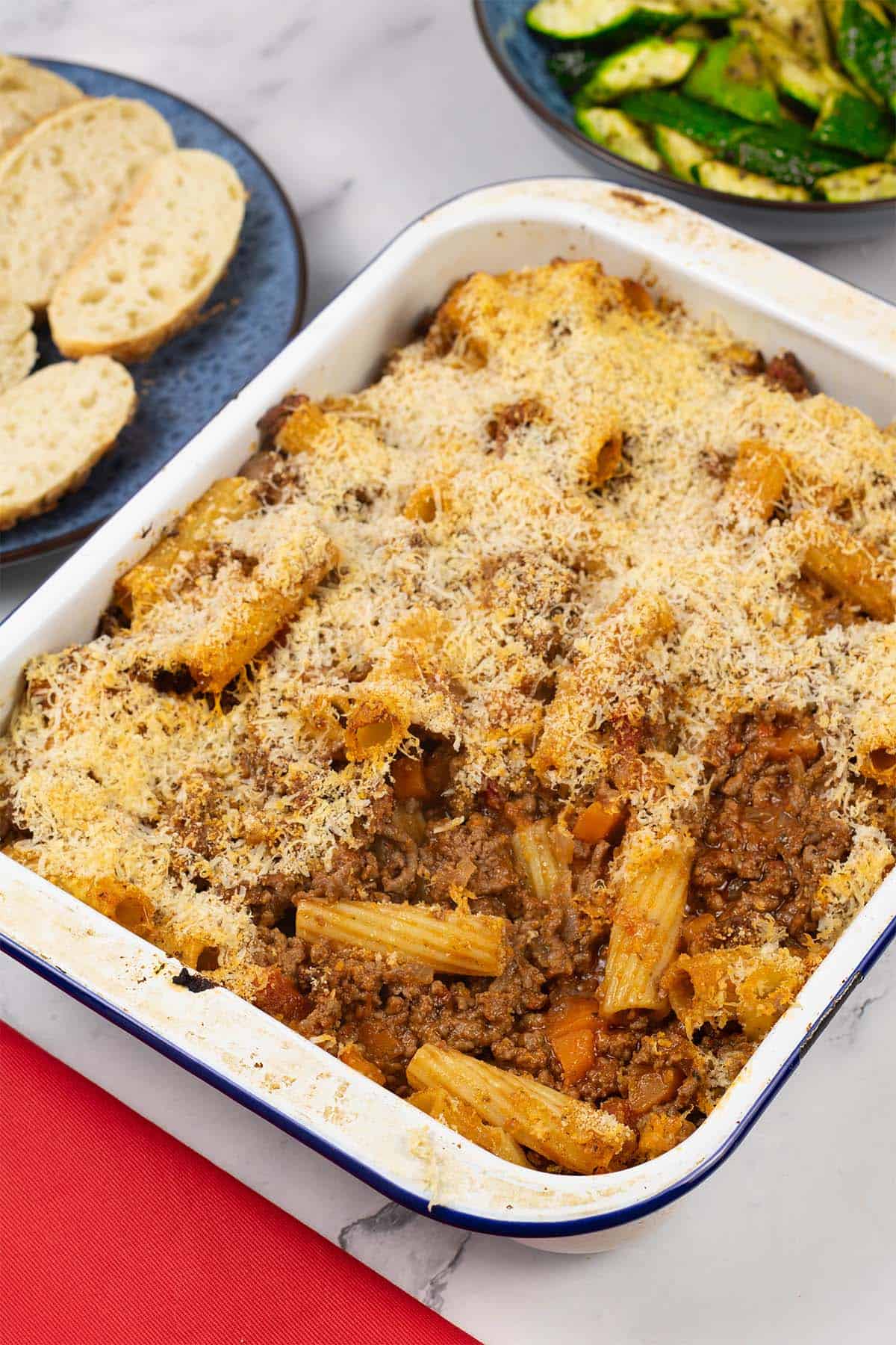 Beef and tomato pasta bake in a blue rimmed white enamel roasting tin with a bowl of air fryer courgettes and plate of sliced crusty bread in the background.