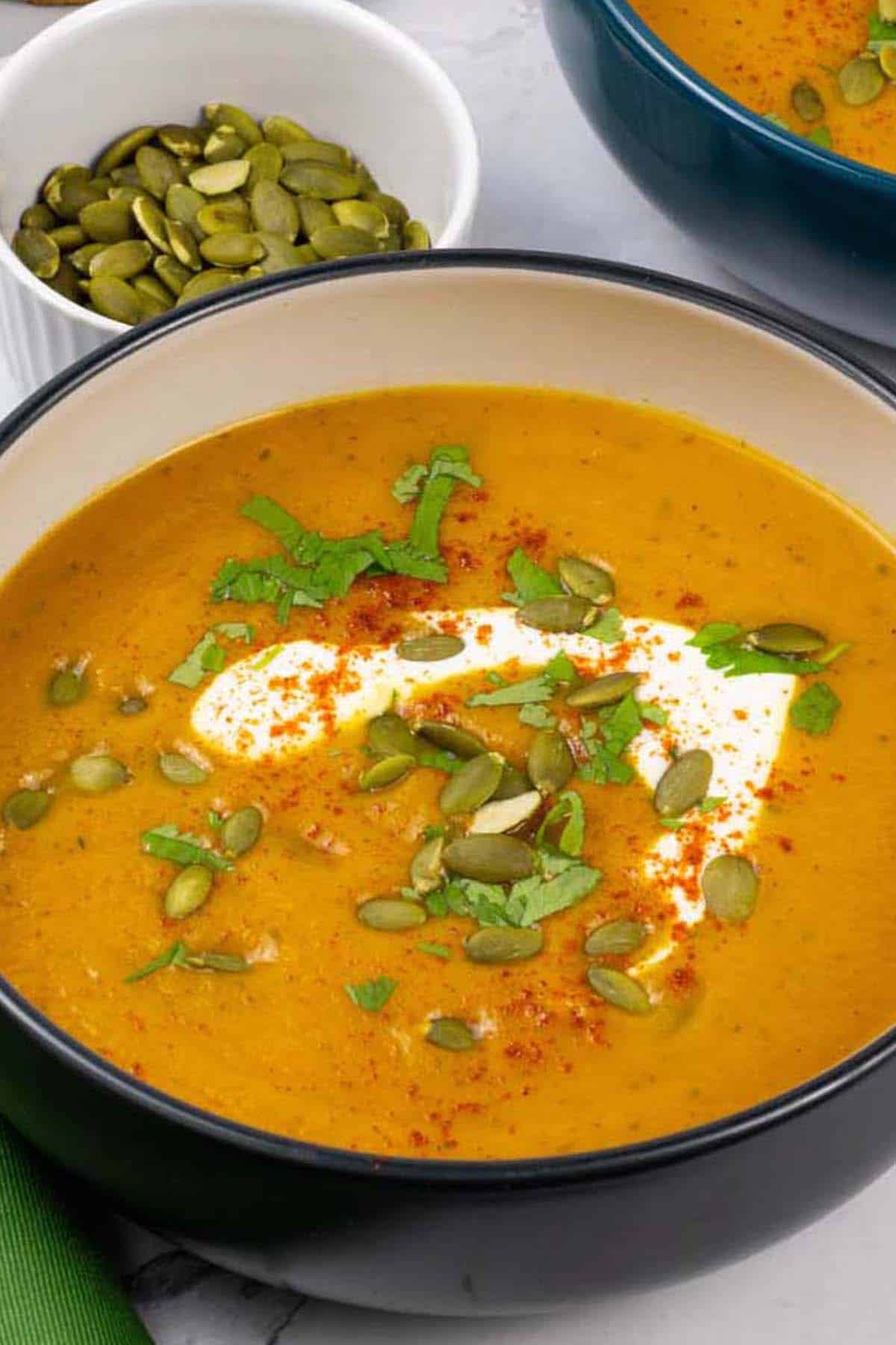 Two bowls of carrot and coriander soup with a small dish of toasted pumpkin seeds in the background.
