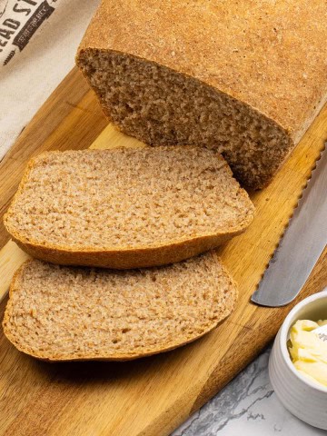 Classic wholemeal bread made in the Thermomix on a board with 2 slices cut and a small dish of butter with butter knife on the side.