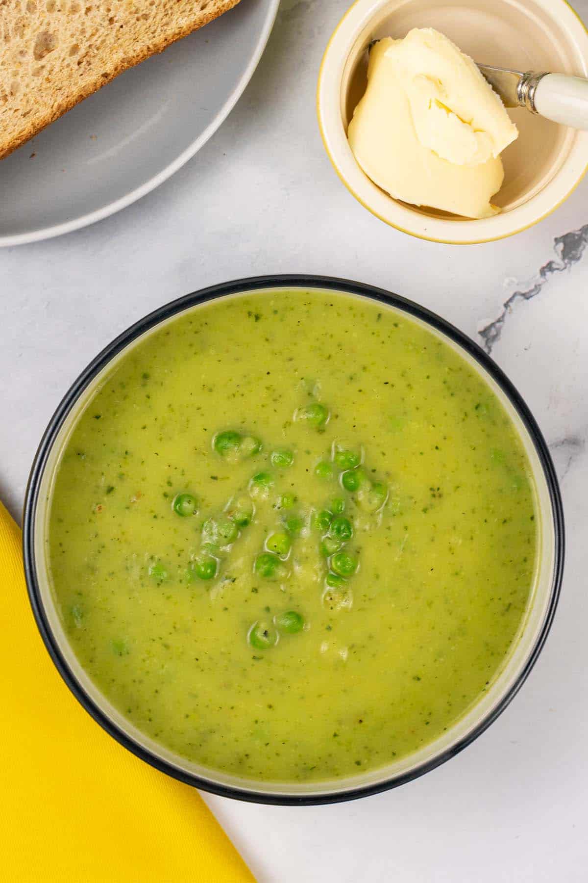Courgette, potato and leek soup in a black and cream bowl with a plate of sliced bread and dish of butter in the background.