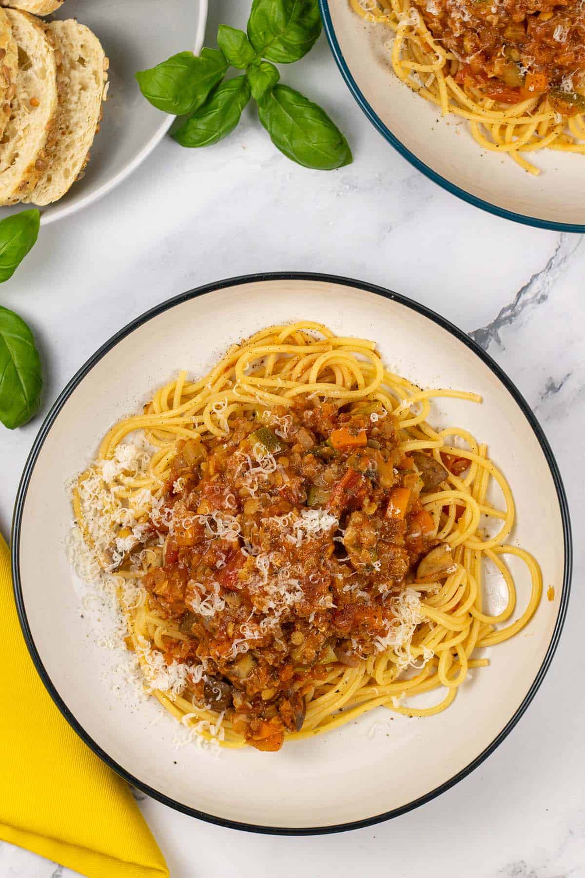 Two bowls of lentil and vegetable bolognese with spaghetti and a plate of sliced bread in the background.