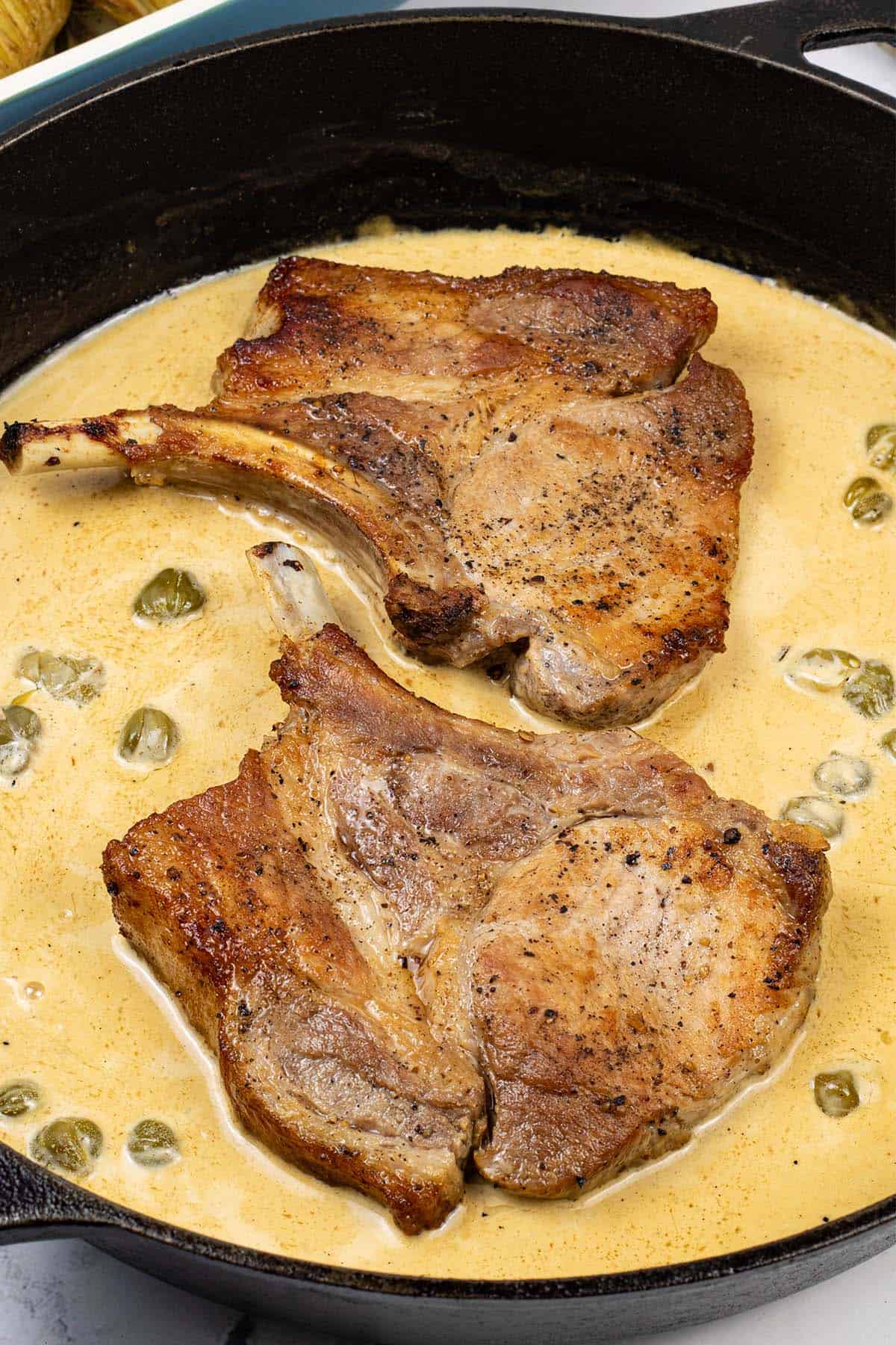 2 portions of pork chops with mustard and capers in a black skillet.