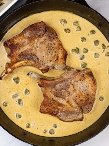2 portions of pork chops with mustard and capers in a black skillet.