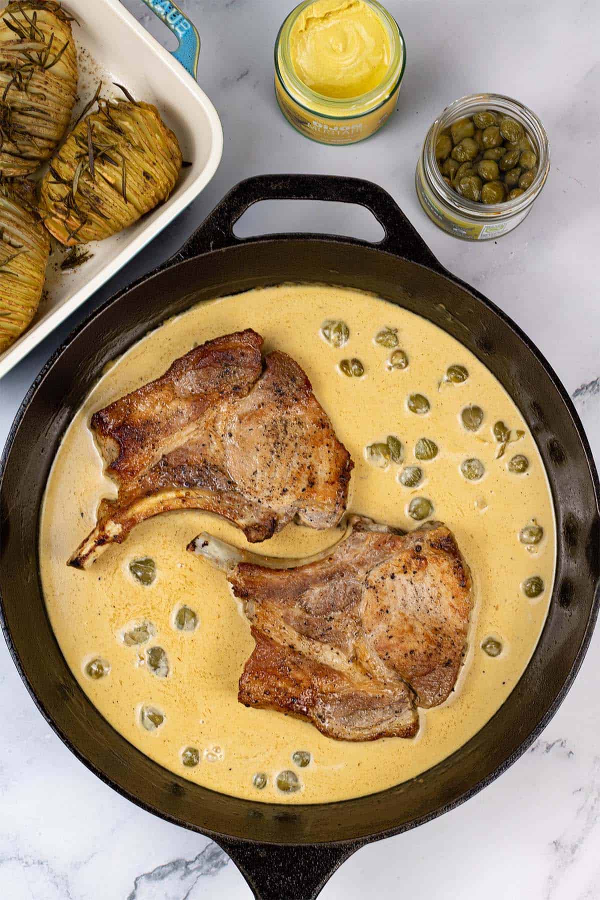 2 portions of pork chops with mustard and capers in a black skillet with garlic and rosemary hasselback potatoes in the background.