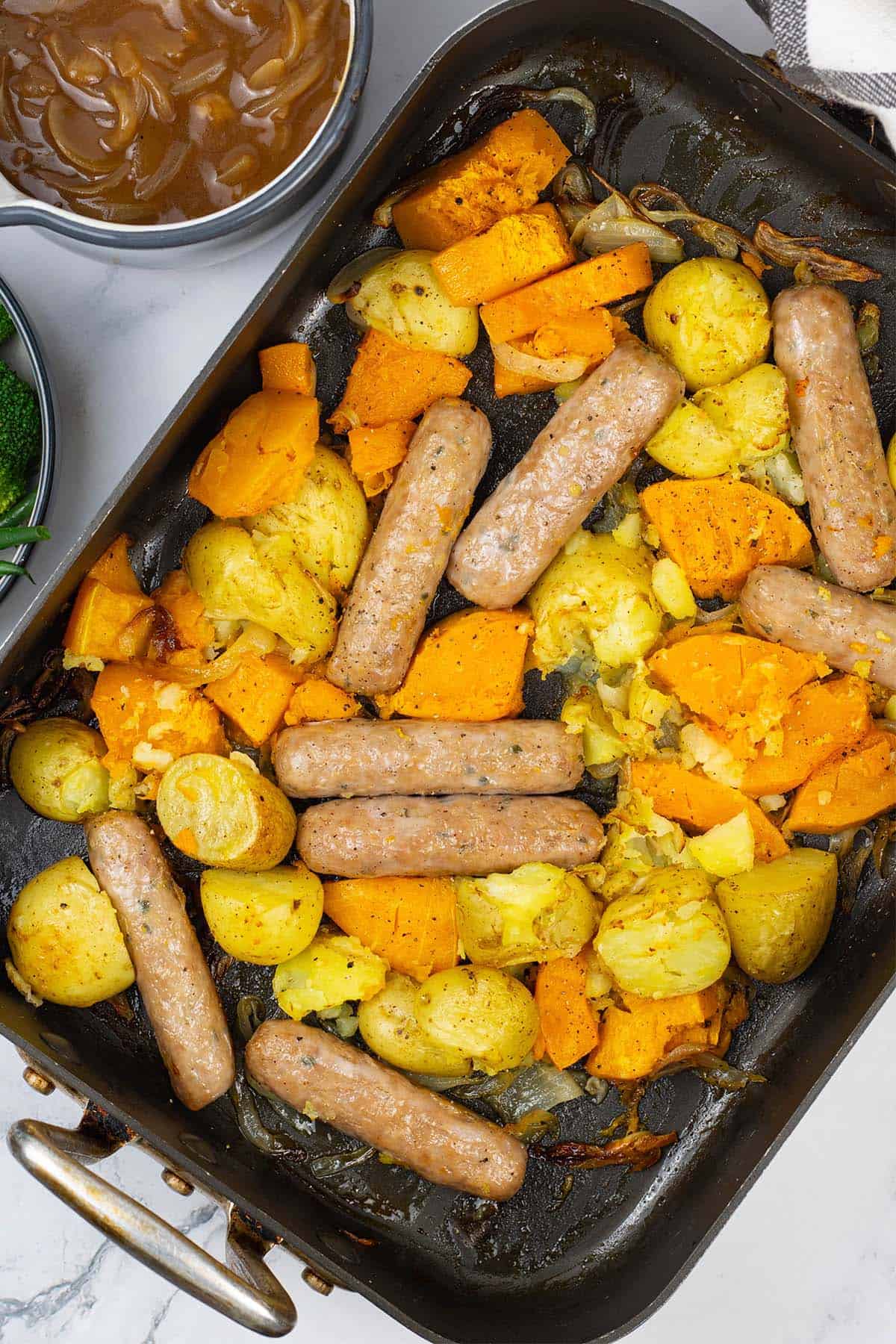 Sausage, squash and potato bake in a large roasting tin with a jug of onion gravy and bowl of broccoli and green beans on the side.