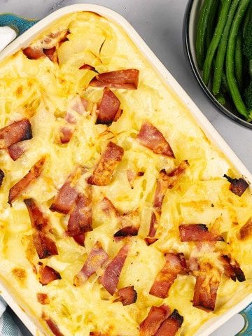 Cheesy potato and gammon bake in a rectangular Staub oven dish with a bowl of green beans and broccoli on the side.