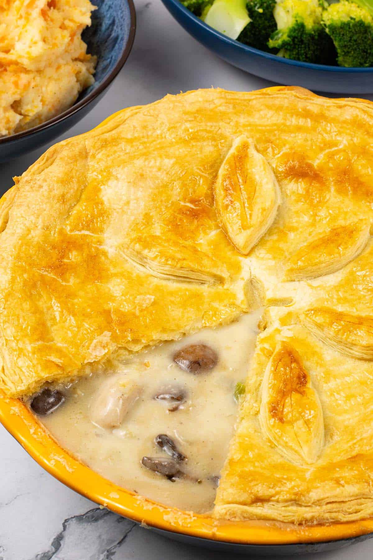 Creamy chicken and mushroom pie in a yellow pie dish with bowls of root veg mash and broccoli in the background.