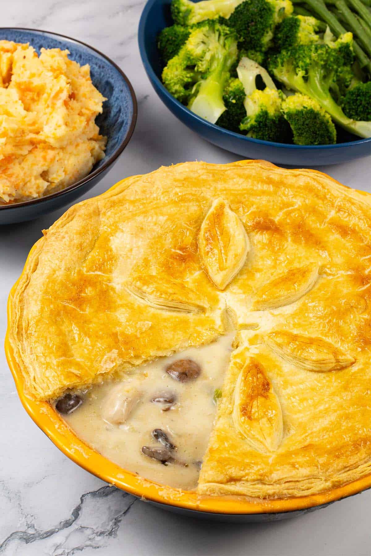 Creamy chicken and mushroom pie in a yellow pie dish with bowls of root veg mash and broccoli in the background.