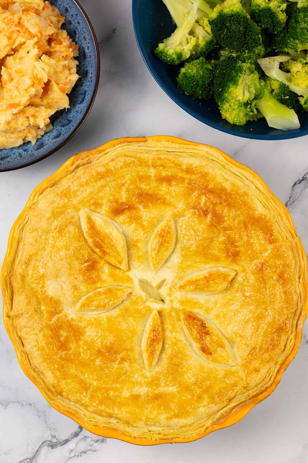 Creamy chicken and mushroom pie in a yellow pie dish with bowls of root veg mash and broccoli in the background.
