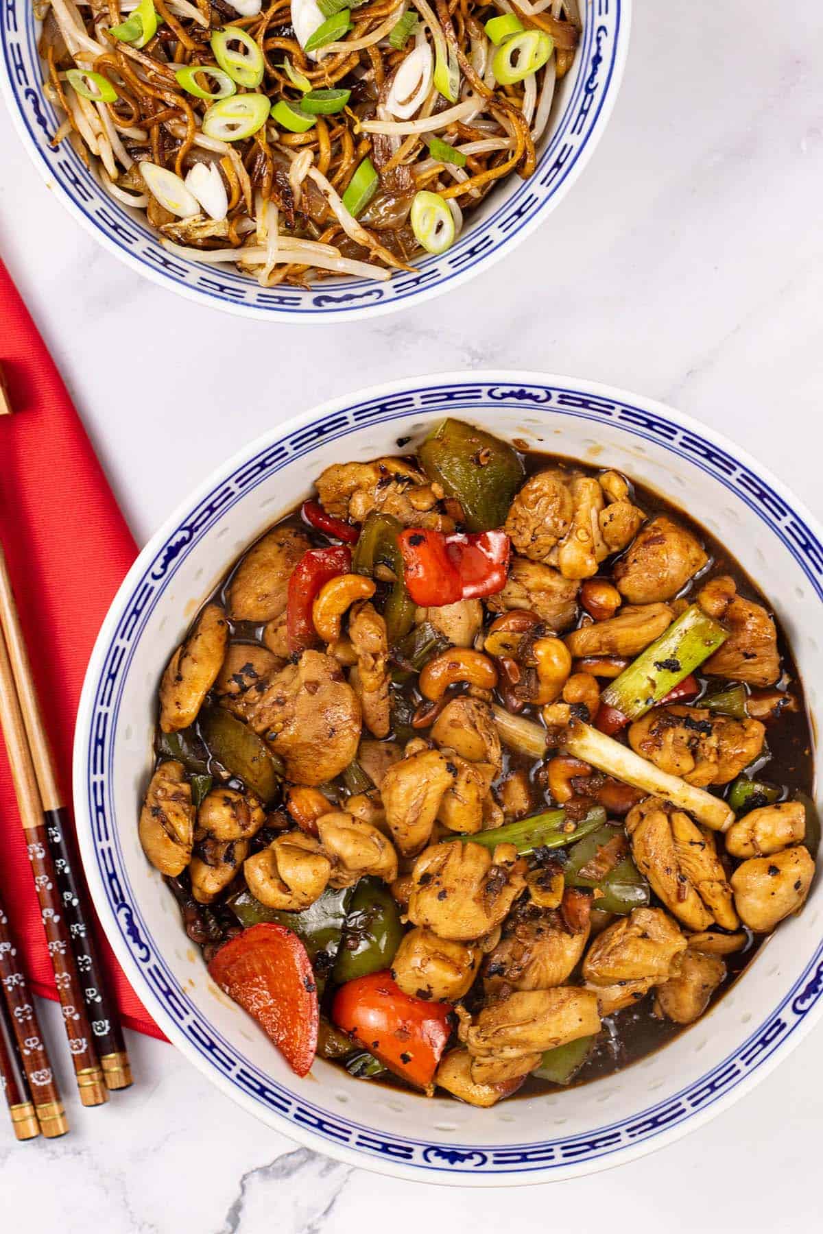 Kung pao chicken in a large blue and white oriental serving bowl with a bowl of fried noodles with beansprouts in the background.