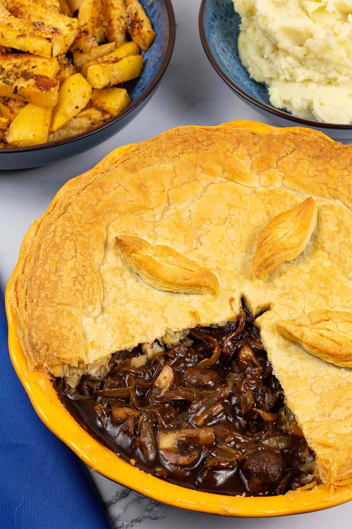 Mushroom and Guinness pie in a round yellow pie dish with bowls of parmesan and rosemary swede, and mashed potatoes on the side.