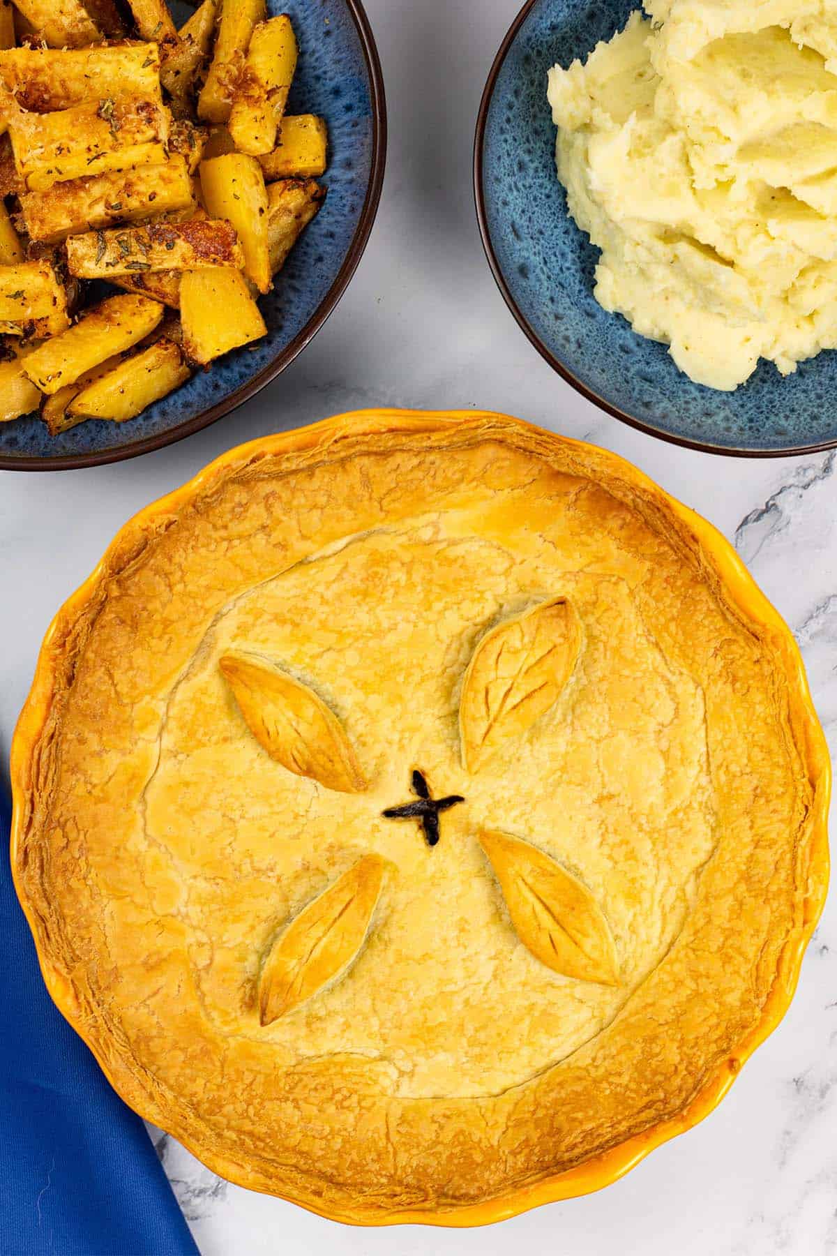 Mushroom and Guinness pie in a round yellow pie dish with bowls of parmesan and rosemary swede, and mashed potatoes on the side.