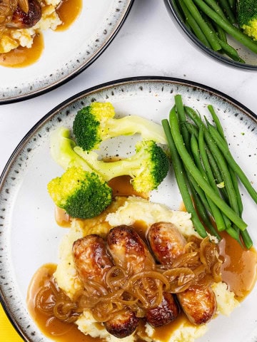 Two plates of sausages with caramelised onion gravy on a bed of mashed potatoes with broccoli and green beans on the side.