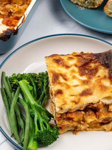 A portion of vegetable lasagne in a teal and cream bowl with tenderstem broccoli and green beans on the side.
