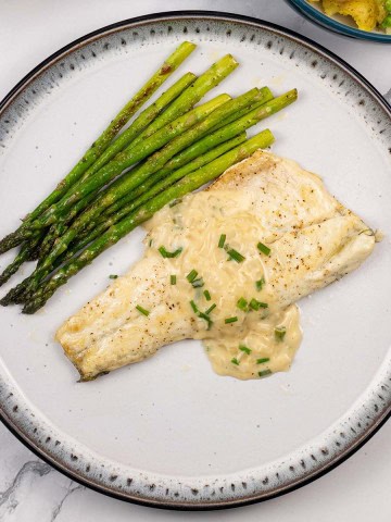 A portion of easy sea bass with white wine and cream sauce on a black and light grey patterned dinner plate with sautéed asparagus.