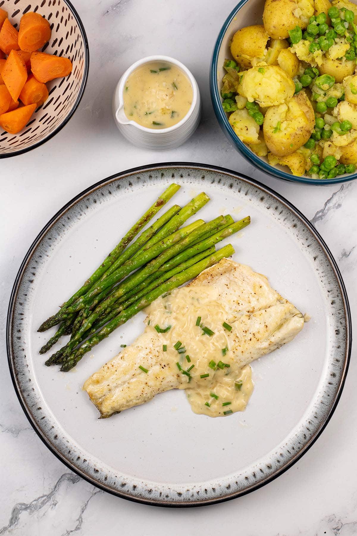 A portion of easy sea bass with white wine and cream sauce on a black and light grey patterned dinner plate with sautéed asparagus.