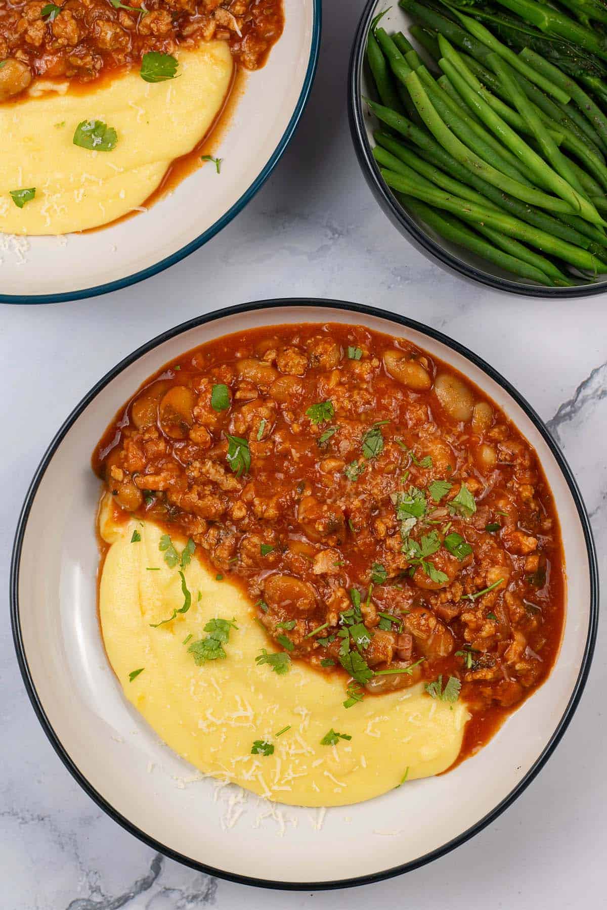 Two bowls of sausage and white bean ragu with cheesy polenta and a bowl of green beans and broccoli on the side.