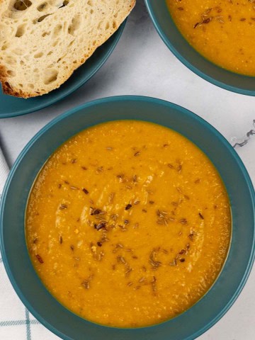 Two bowls of spiced carrot and lentil soup with a plate of sliced crusty bread on the side.