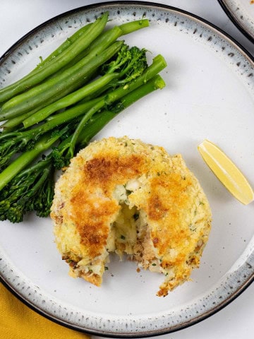 A melt in the middle fishcake on a black and white patterned dinner plate with green beans and tenderstem broccoli, and a wedge of lemon.