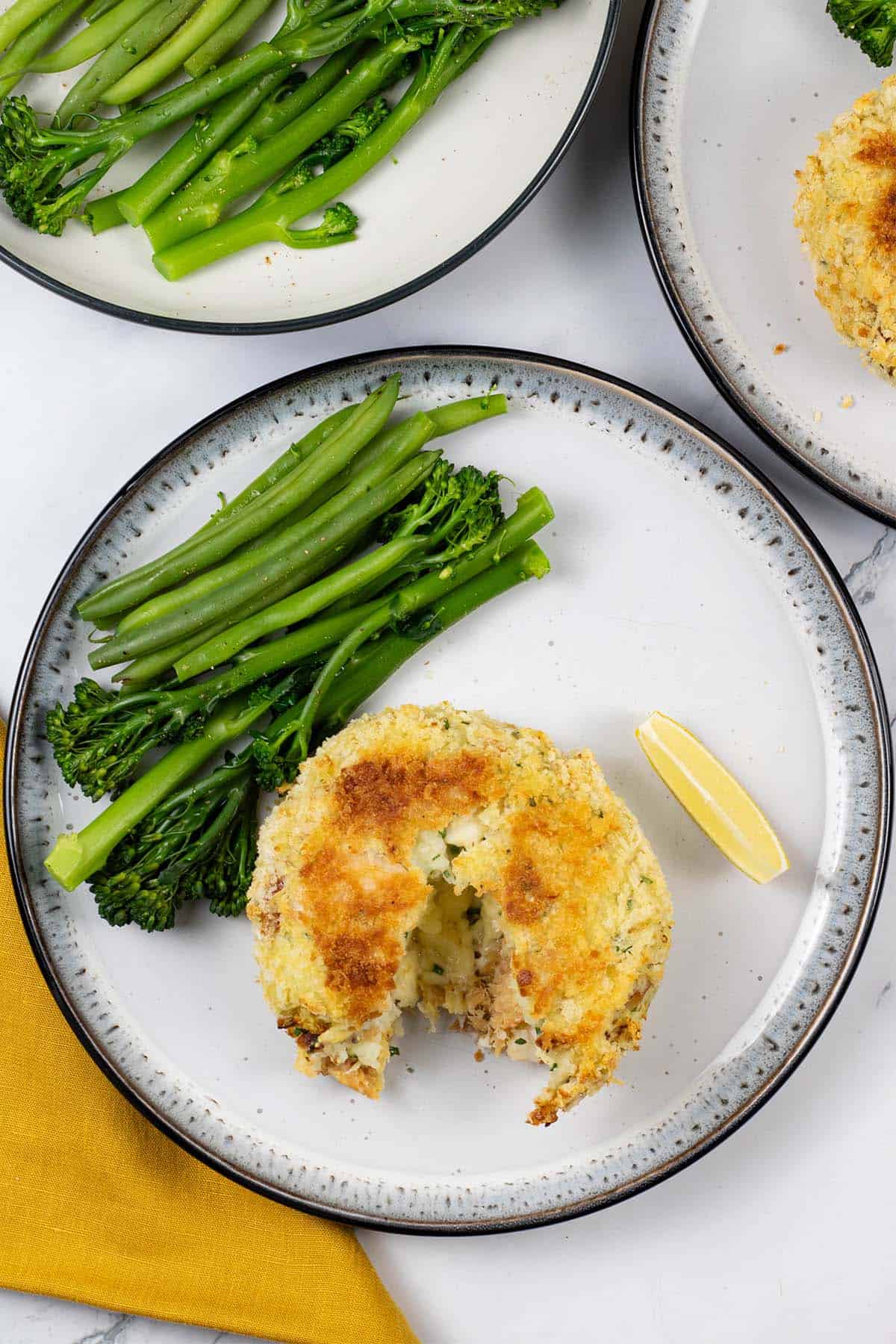 A melt in the middle fishcake on a black and white patterned dinner plate with green beans and tenderstem broccoli, and a wedge of lemon.
