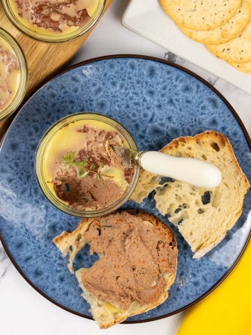 A ramekin of chicken liver pate sitting on a blue patterned side plate with toasted crusty bread. Ramekin dishes of pate and a plate of crackers in the background.