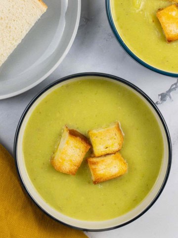 Two bowls of leek and potato soup garnished with croutons and a plate of sliced bread on the side.