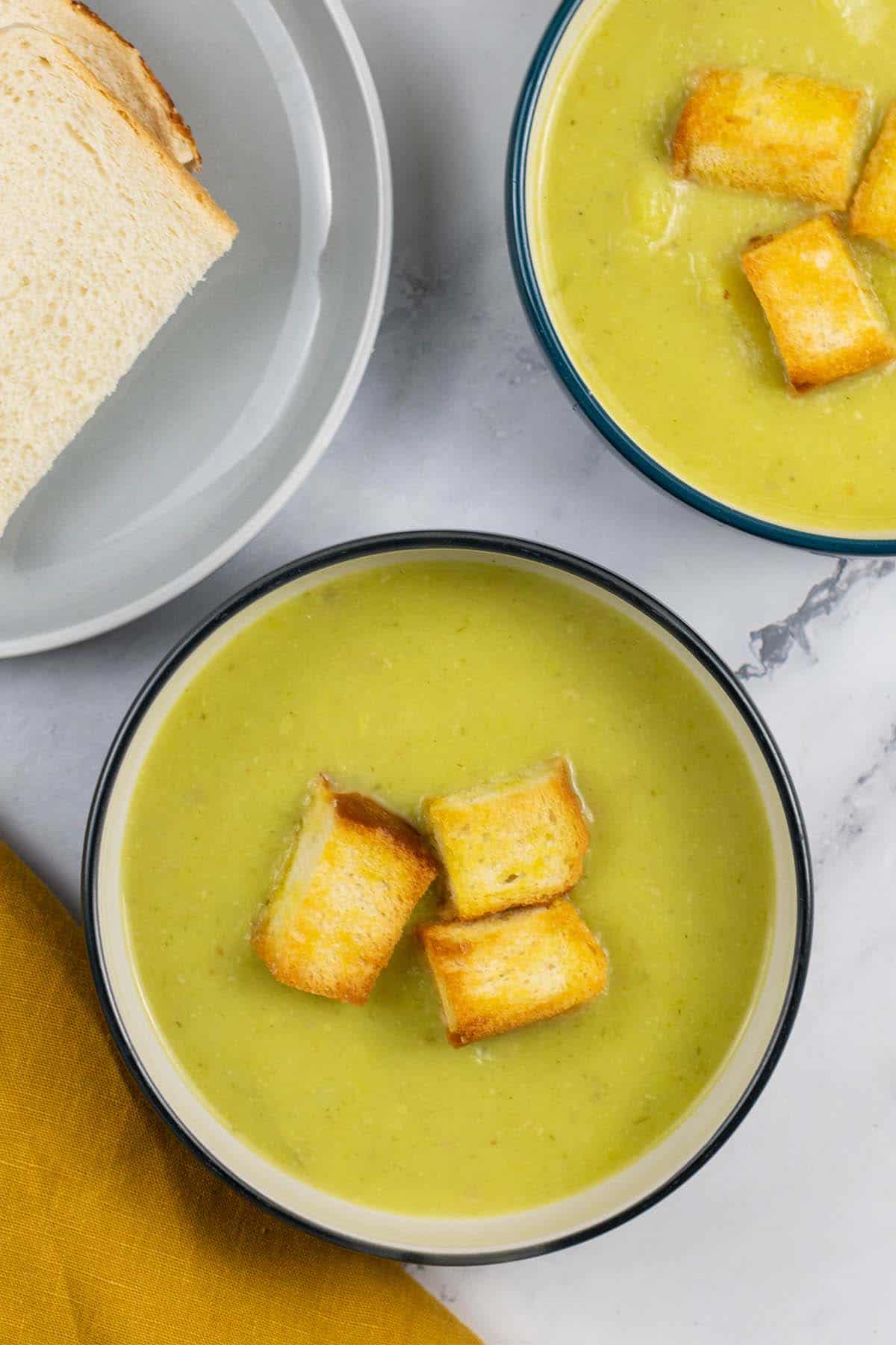 Two bowls of leek and potato soup garnished with croutons and a plate of sliced bread on the side.