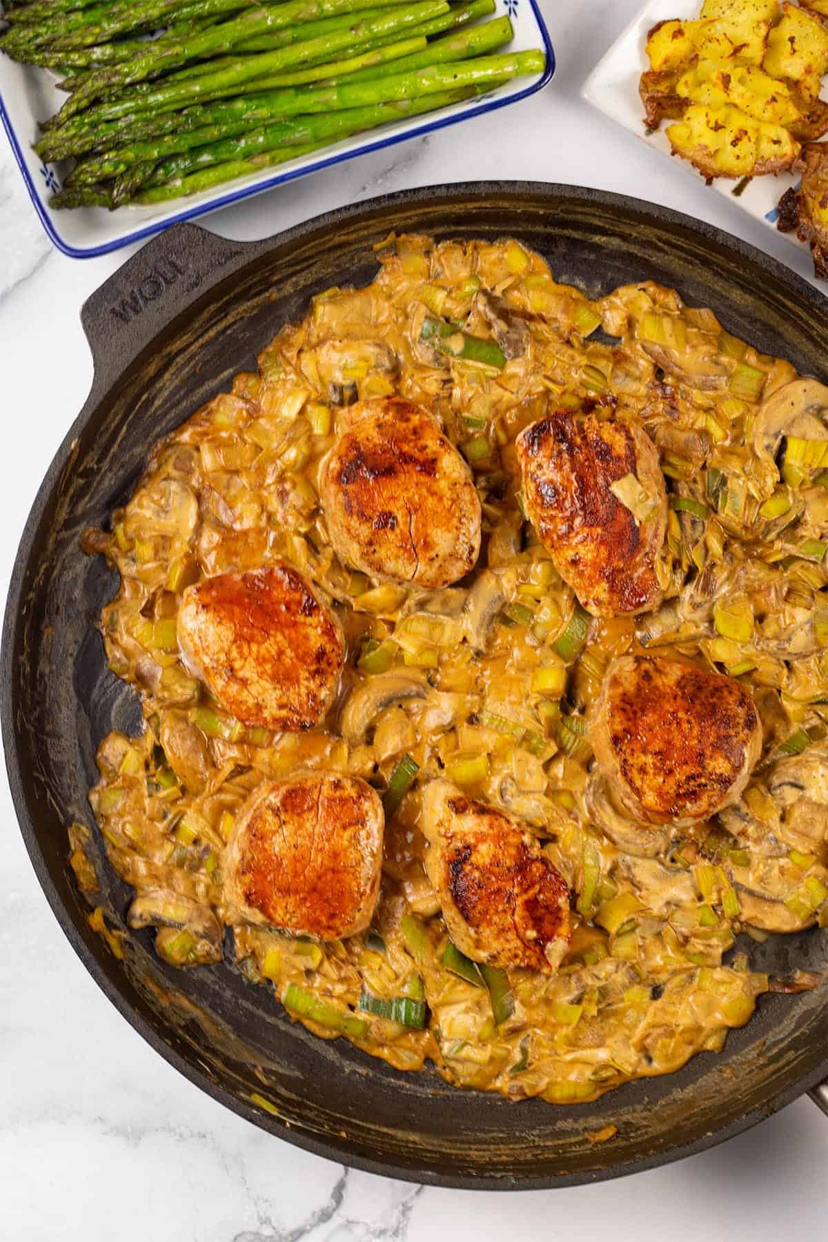 Pork fillet with mushroom and leek sauce in a large frying pan with portions of roast smashed baby potatoes and sauteed asaparagus on the side.
