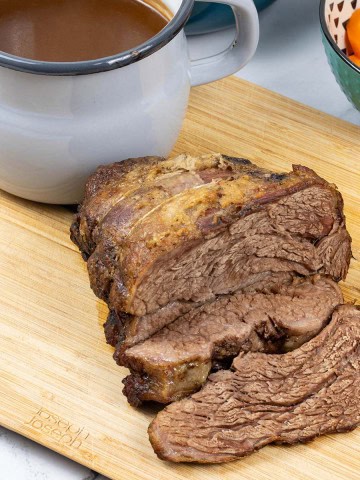 Slow cooker beef pot roast sitting on wooden chopping board with jug of gravy and a bowl of carrots on the side.