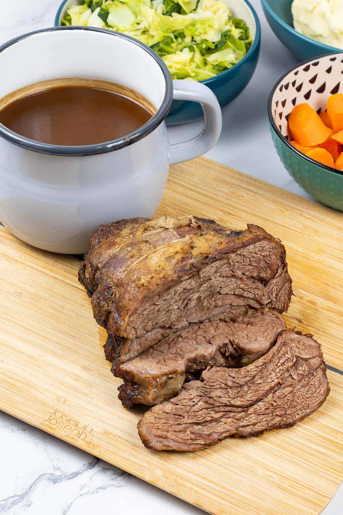 Slow cooker beef pot roast sitting on wooden chopping board with jug of gravy and bowls of cabbage, carrots and mash on the side.