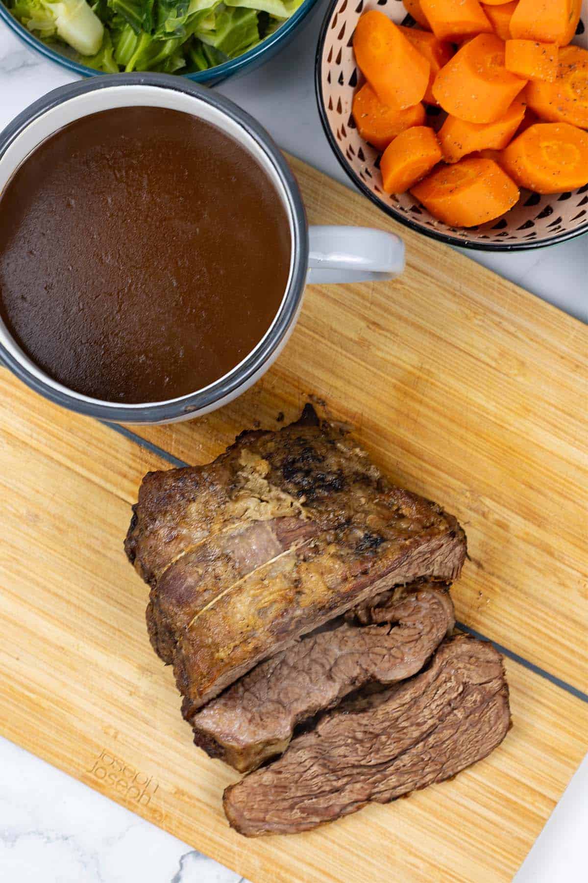 Slow cooker beef pot roast sitting on wooden chopping board with jug of gravy and bowls of cabbage and carrots on the side.