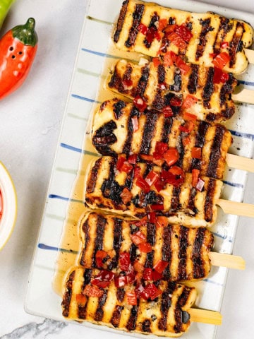 Sweet chilli halloumi skewers on a striped platter with a small dish of sauce on the side.
