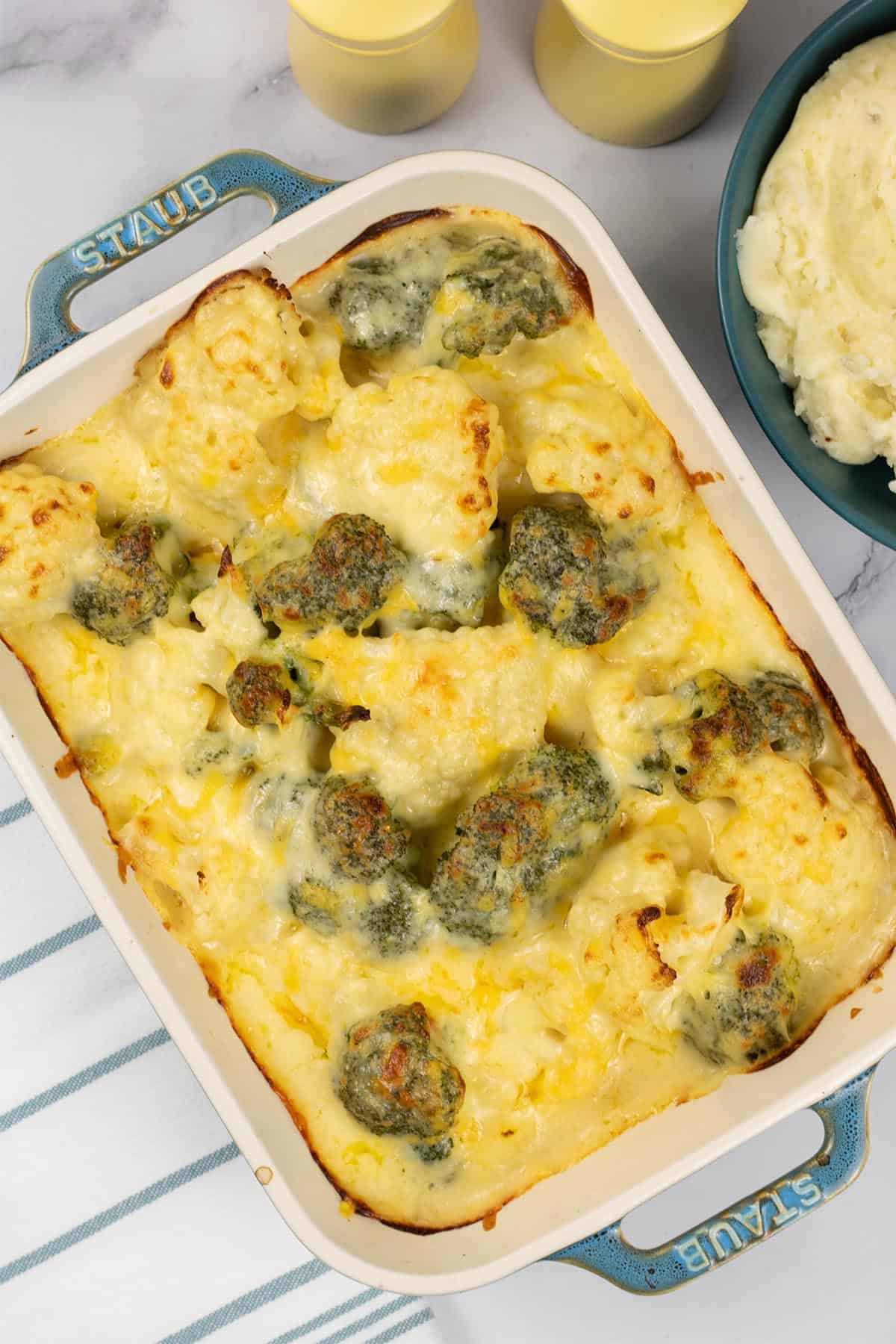 Cauliflower and broccoli cheese in a rectangle Staub oven dish with a bowl of mashed potatoes on the side.