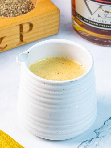 Creamy peppercorn sauce in a small ribbed white jug with a pot of crushed peppercorns and bottle of brandy in the background.