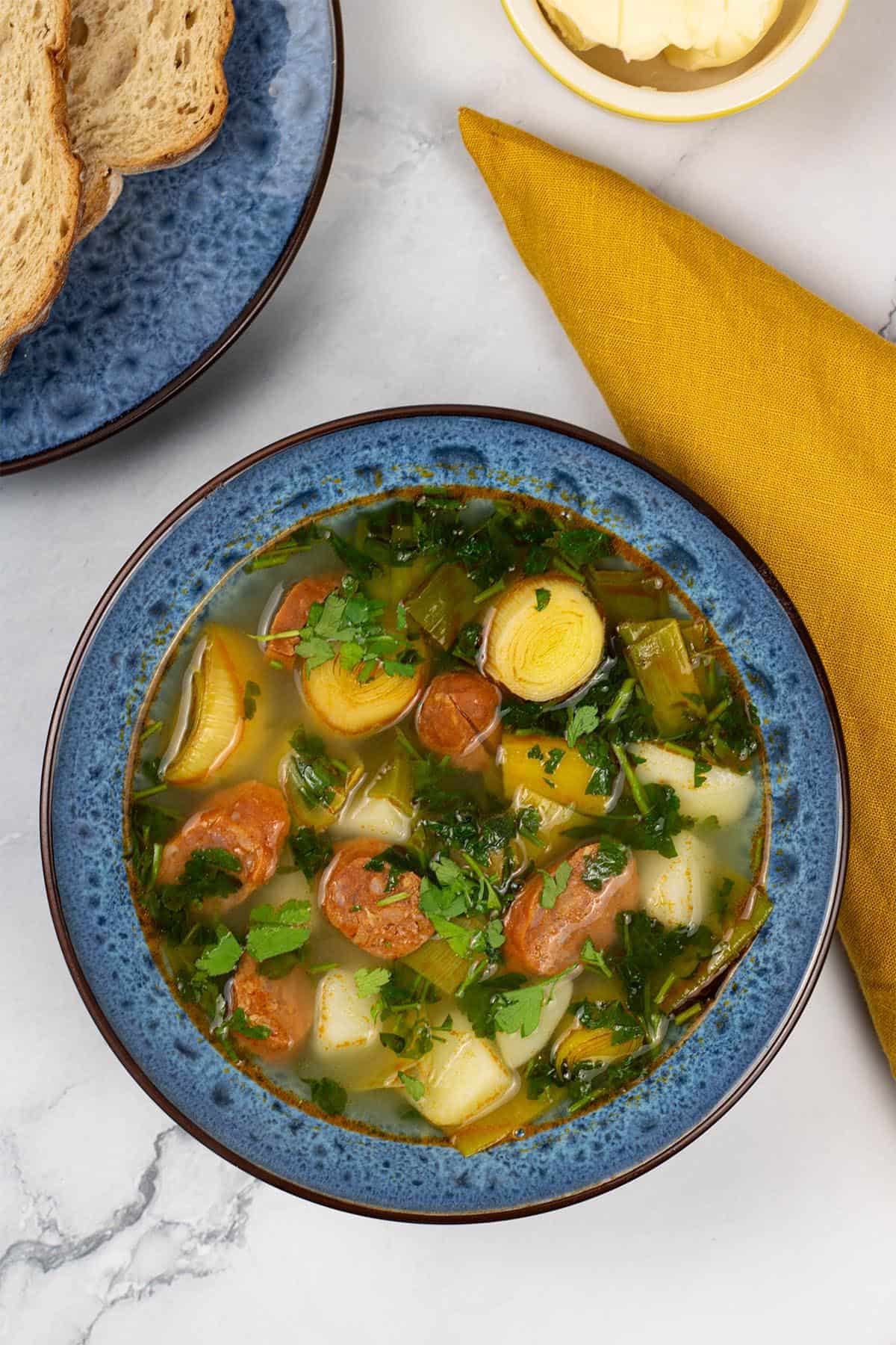 A portion of potato, leek and chorizo soup in a blue patterned bowl with a plate of bread, small dish of butter and mustard napkin on the side.