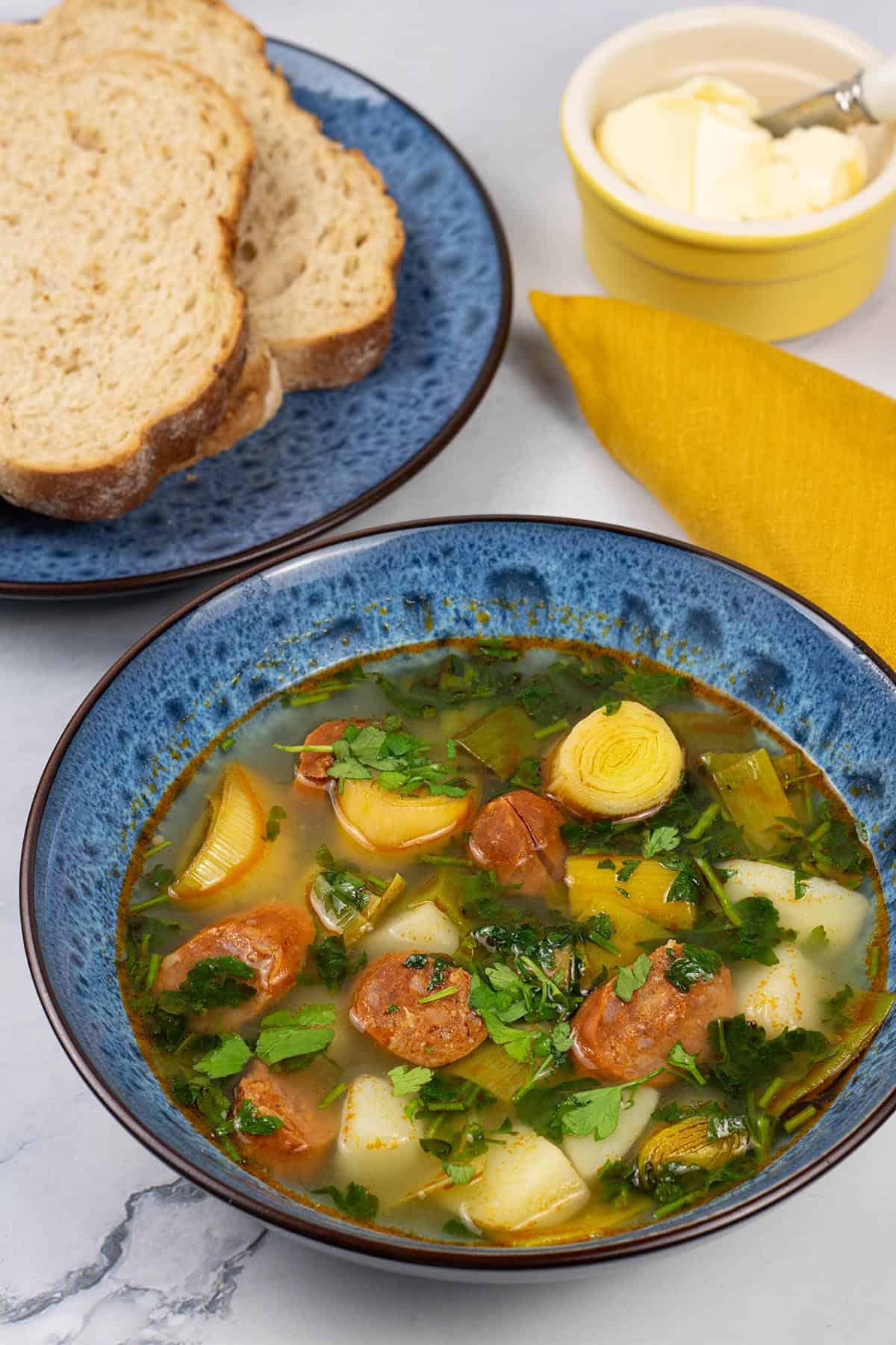 A portion of potato, leek and chorizo soup in a blue patterned bowl with a plate of bread, small dish of butter and mustard napkin on the side.